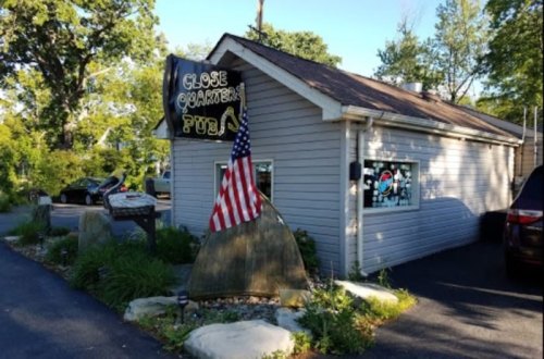 A small pub with a sign reading "Close Quarters Pub," featuring an American flag and surrounded by greenery.