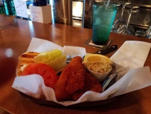 A plate with fried fish, lettuce, tomato, coleslaw, a lemon wedge, and a grilled sandwich on a wooden table.