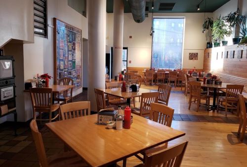 A spacious, empty restaurant with wooden tables and chairs, featuring plants and a large window.