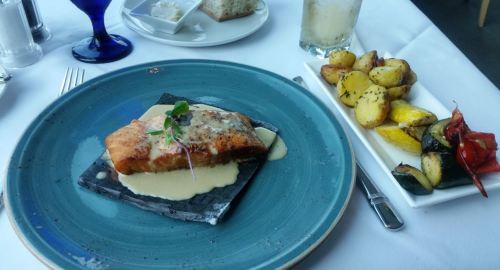 A plate of salmon with sauce on a slate, accompanied by roasted potatoes and grilled vegetables on the side.