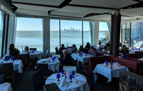 A restaurant with large windows overlooking a lake and city skyline, featuring tables set with blue glasses and diners.