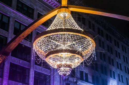 A large, sparkling chandelier hangs from a structure, illuminated against a dark city backdrop.
