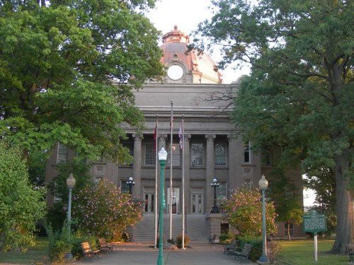 Historic building with a dome, surrounded by trees and benches, featuring flags in front and a well-maintained lawn.