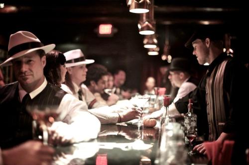 A vintage-style bar scene with patrons in 1920s attire, enjoying drinks and socializing under dim lighting.