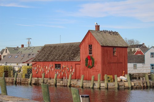 A red wooden building with a wreath, situated by a calm waterway, surrounded by wooden posts and a clear blue sky.