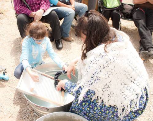 A woman and a girl wash clothes together in a basin, surrounded by people sitting nearby.