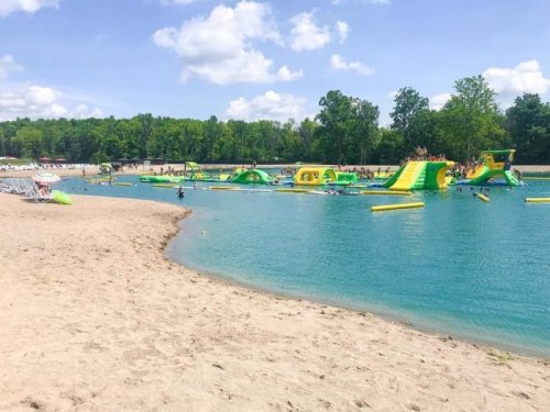 A sandy beach with a clear blue lake featuring inflatable water attractions and people enjoying the sun.