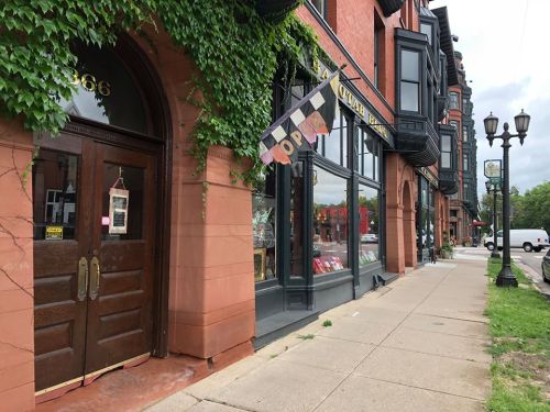 A brick building with ivy, featuring an "Open" sign and colorful flags, along a sidewalk on a cloudy day.