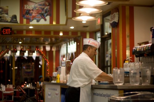 A diner scene with a man in a white shirt and hat preparing drinks at the counter, surrounded by retro decor.