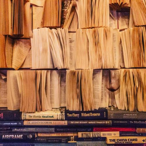 A wall of books with pages fanned out, featuring a wine glass resting on a stack of novels at the bottom.