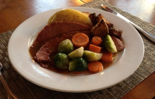 A plate of sliced meat with carrots, Brussels sprouts, potatoes, and roasted vegetables, served on a wooden table.