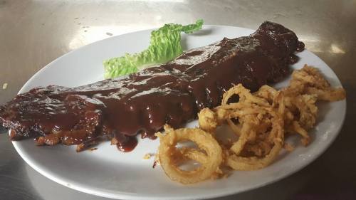 A plate featuring a large BBQ rib covered in sauce, accompanied by crispy onion rings and a piece of lettuce.