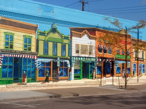 Colorful mural depicting historic storefronts with people walking along a street, showcasing vibrant urban art.