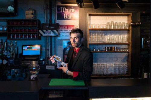 A magician in a black suit holds playing cards at a bar, with shelves of glasses and bottles in the background.
