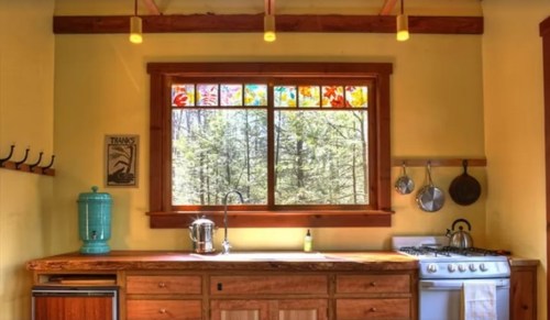 A cozy kitchen with wooden cabinets, a colorful stained glass window, and a view of trees outside.