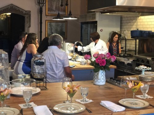 A chef demonstrates cooking techniques in a stylish kitchen while guests watch, surrounded by elegant table settings.