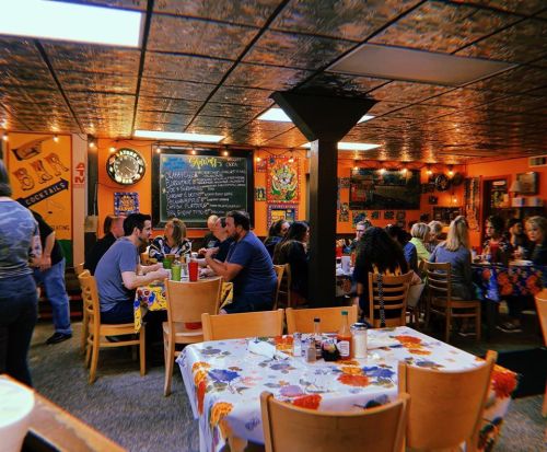 A bustling restaurant interior with diners at tables, colorful decor, and a chalkboard menu on the wall.