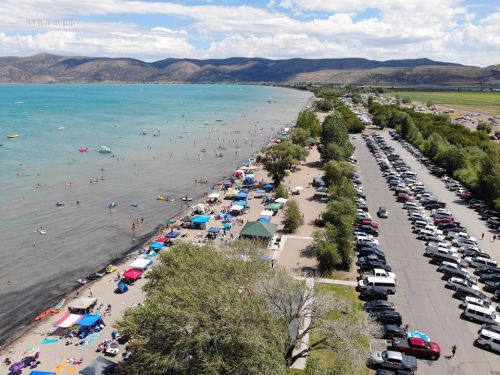 Aerial view of a beach with people, colorful umbrellas, and parked cars along the shore, surrounded by mountains.