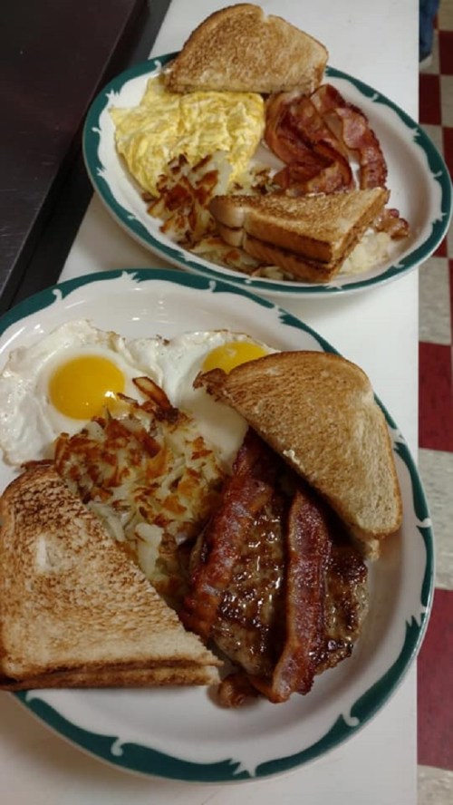 Two plates of breakfast featuring scrambled eggs, bacon, hash browns, toast, and sausage.