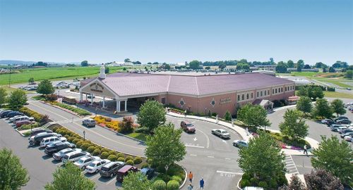 Aerial view of a large building with a red roof, surrounded by greenery and parked cars in a spacious lot.