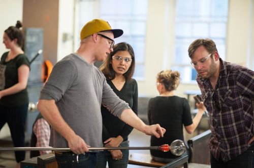 A glassblowing instructor demonstrates technique to two students in a workshop setting. Others are working in the background.