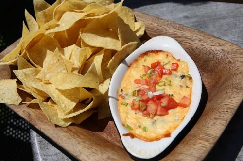 A wooden platter with crispy tortilla chips beside a small dish of cheesy dip topped with diced tomatoes and green onions.