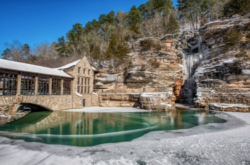 A scenic view of a stone building by a frozen pond, with a waterfall cascading down a rocky cliff in winter.