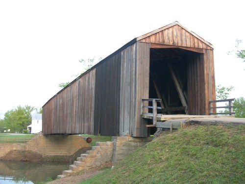 A wooden covered bridge spans a small waterway, with grassy banks and a clear sky in the background.