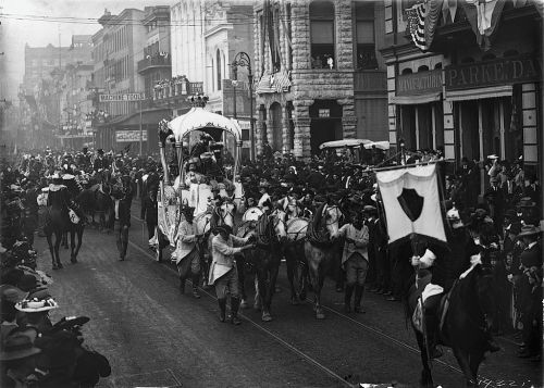 A historic parade scene with a horse-drawn carriage, crowds, and banners lining the street.