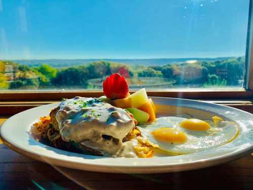 A plate of breakfast with eggs, hash browns, and fruit, set against a scenic view of rolling hills and blue sky.