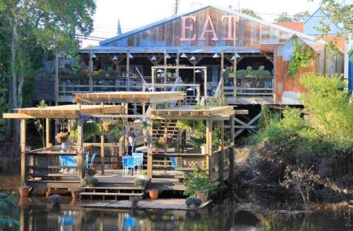 A rustic restaurant by the water with a sign reading "EAT," featuring outdoor seating and lush greenery.