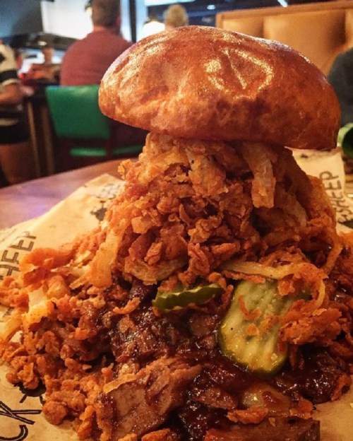 A towering sandwich with crispy fried onions, pickles, and barbecue sauce on a soft bun, served on a paper-lined plate.