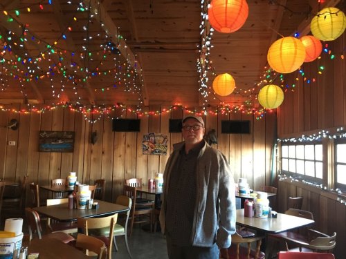 A man stands in a cozy restaurant with wooden walls, colorful lanterns, and festive string lights.