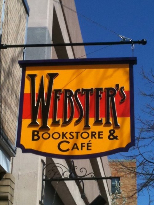 Sign for Webster's Bookstore & Café, featuring bold lettering on a bright yellow background against a clear blue sky.