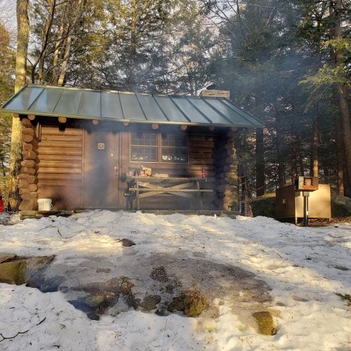 A cozy log cabin surrounded by snow, with smoke rising from a chimney and trees in the background.