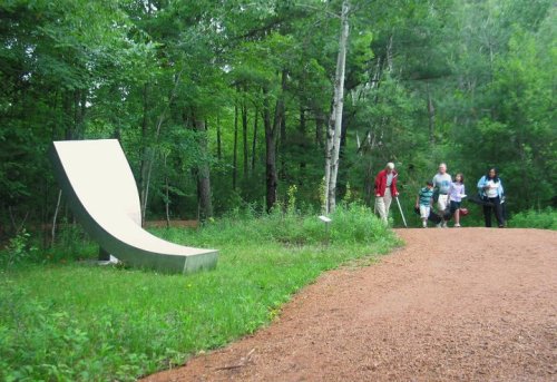 A curved concrete sculpture beside a walking path in a green forest, with people strolling nearby.