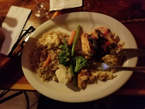 A plate of grilled chicken with vegetables, rice, and a side of mixed veggies, served on a wooden table.