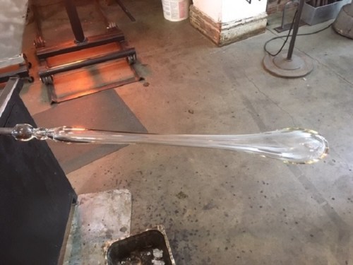 A long, clear glass object being shaped in a glassblowing workshop, with tools and equipment in the background.