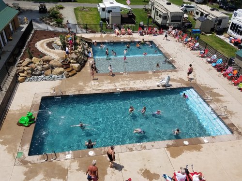 Aerial view of a busy pool area with two pools, people swimming, and lounge chairs around, set in a campground.