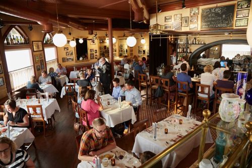A bustling restaurant interior with diners at tables, a bar area, and vintage decor on the walls.