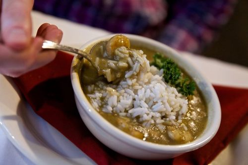 A bowl of gumbo with rice, garnished with parsley, on a white plate with a red napkin. A hand holds a spoon.