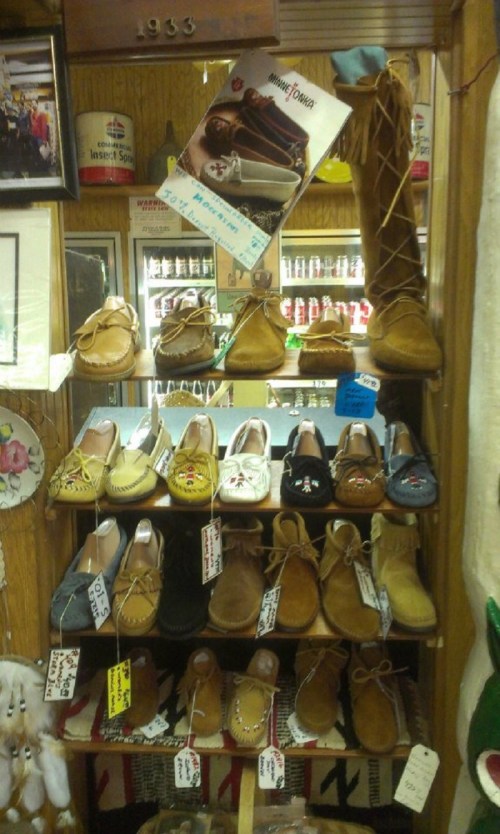 A display of various styles of moccasins and slippers on shelves, with price tags and a promotional poster above.