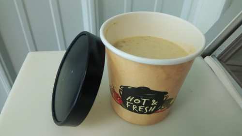 A cup of creamy soup with a black lid, labeled "Hot & Fresh," placed on a light surface.