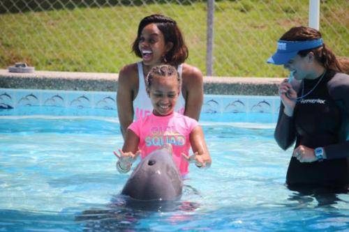 A girl smiles joyfully as she interacts with a dolphin in a pool, accompanied by two women.
