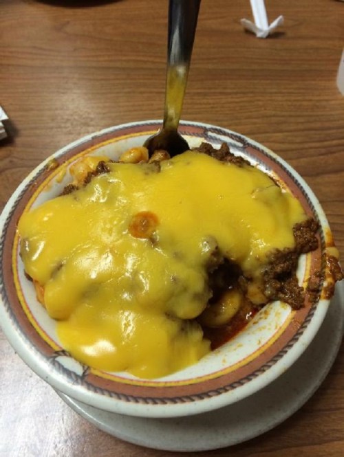 A bowl of chili topped with melted cheese, served with a spoon, on a wooden table.