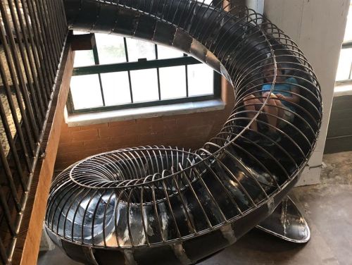 A spiral metal slide inside a building, with a person visible at the top, surrounded by large windows.