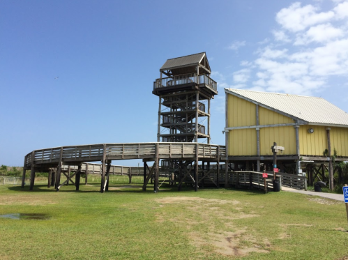 A wooden observation tower next to a yellow building, set against a clear blue sky and green grass.