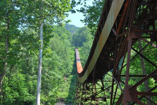 A long, rusted bridge stretches over a lush green forest, surrounded by trees and hills under a clear blue sky.