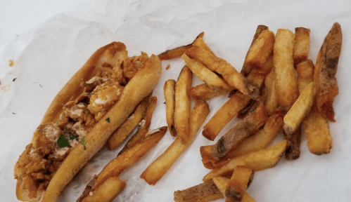 A sandwich filled with fried chicken and herbs, served with a side of golden, crispy French fries on white paper.