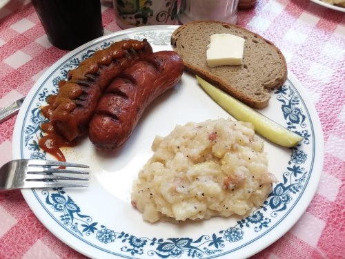 A plate with two sausages, a slice of bread with butter, a scoop of creamy potato dish, and a slice of apple.